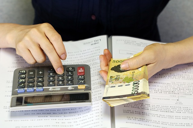 *Property Investment Strategies*—A person using a calculator while holding a stack of 50,000 Korean won banknotes over an open book, symbolizing financial planning, real estate investment calculations, and strategic decision-making.