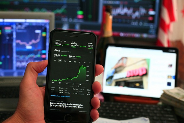 Person analyzing GME stock data on a smartphone with multiple monitors displaying charts in the background, representing real-time stock market forecast activity.